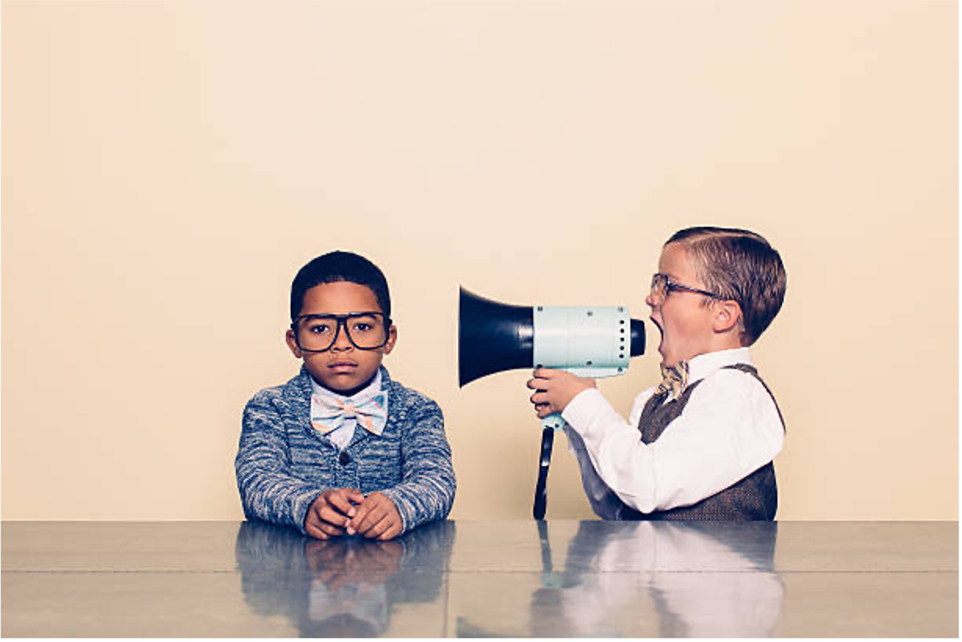 Two children sitting at a table on a yellow background, one of them holding a megaphone