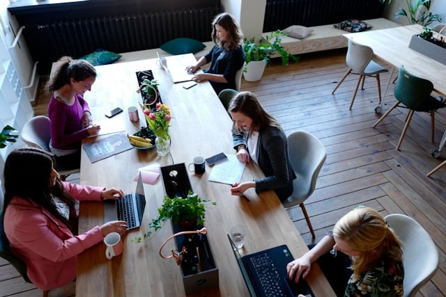 5 women sitting at computers, around a desk